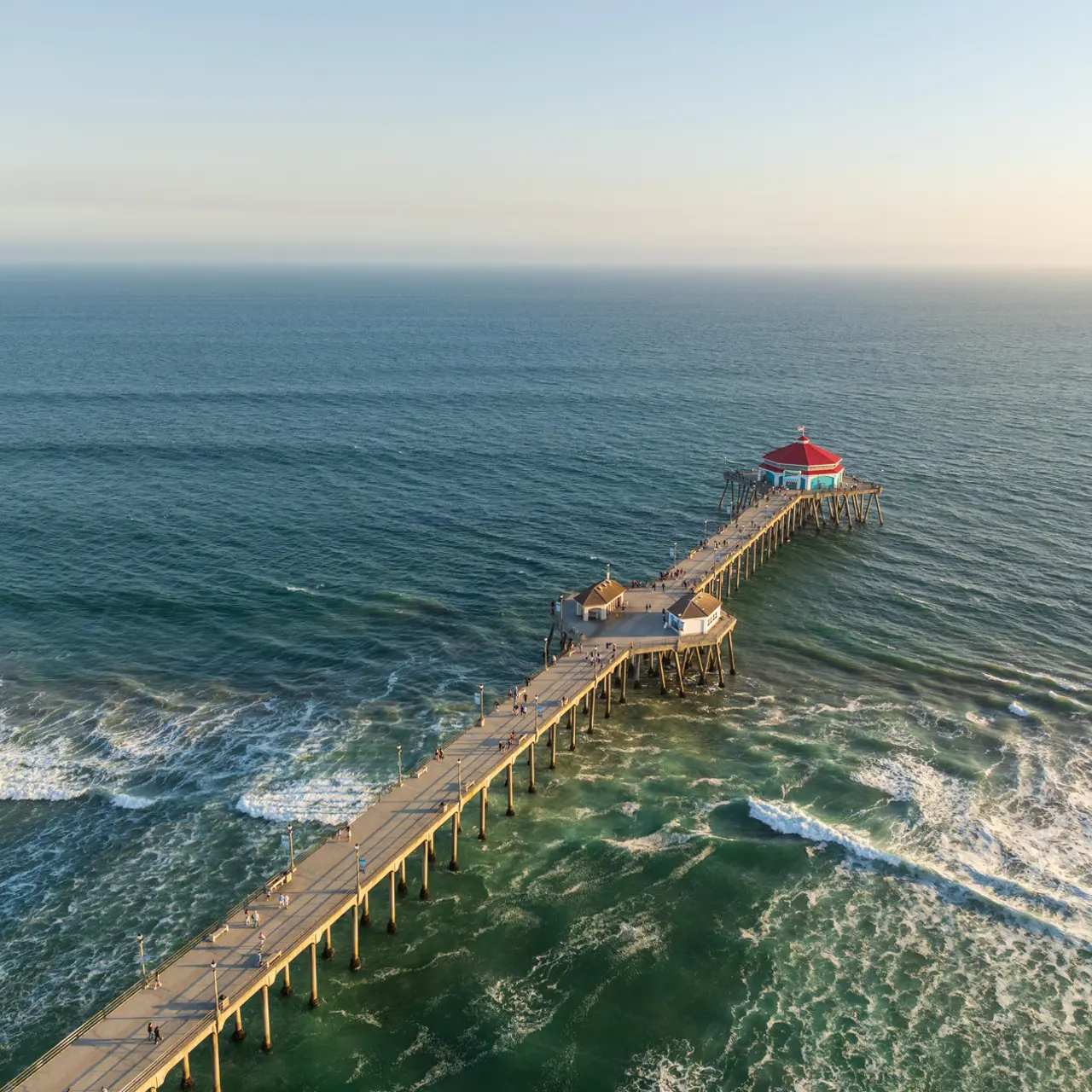 Aerial view of a wooden pier extending into the ocean, with gentle waves crashing against its sides, a small gazebo on the pier, and a red-roofed structure at the end.