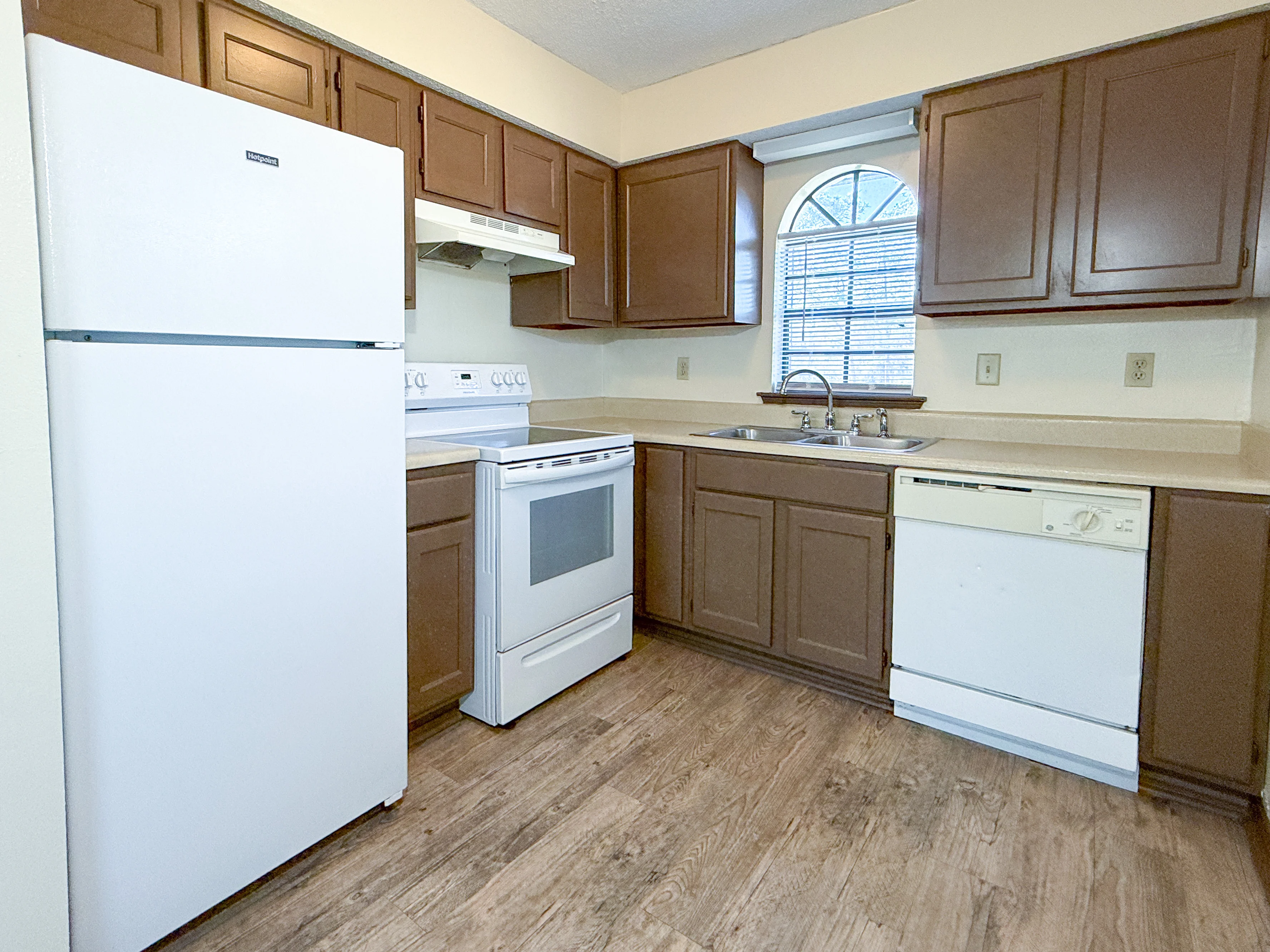 Modern kitchen featuring white appliances, brown cabinets, and a window above the sink, with wooden flooring.