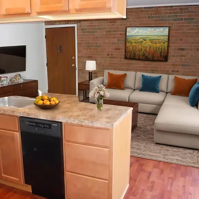 A modern kitchen and living room setup, featuring an open space with a kitchen counter, a black dishwasher, a bowl of oranges, a comfortable sofa with colorful throw pillows, and a TV mounted on the wall.