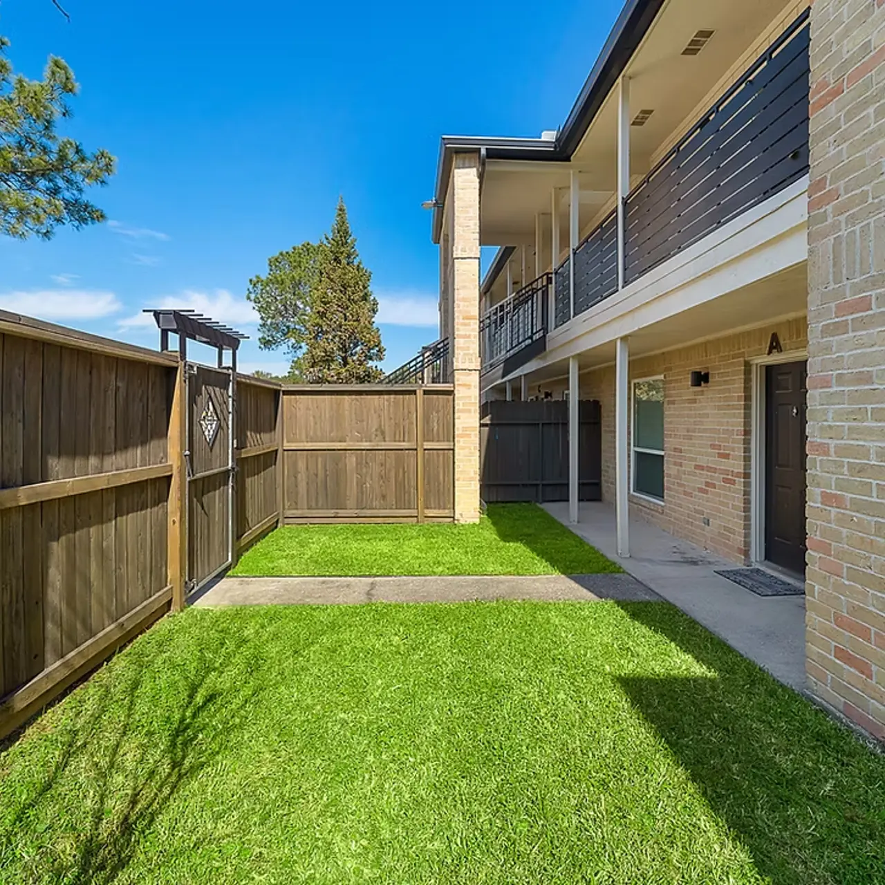 Exterior view of an apartment complex with a small garden area, wooden fencing, and blue skies
