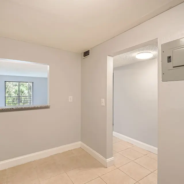 An interior view of a kitchen area with light grey walls, tan tiled flooring, and a silver refrigerator on the left. A small opening leads to another room, and there's a wall-mounted electrical panel.