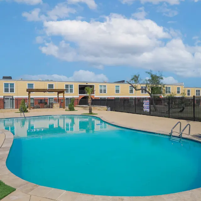 A vibrant swimming pool area surrounded by landscaped greenery and a modern building in the background under a clear blue sky.