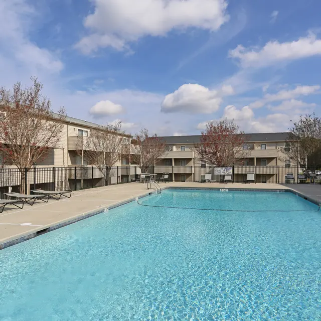 A clear swimming pool surrounded by lounge chairs and two-story apartment buildings under a blue sky with fluffy clouds.
