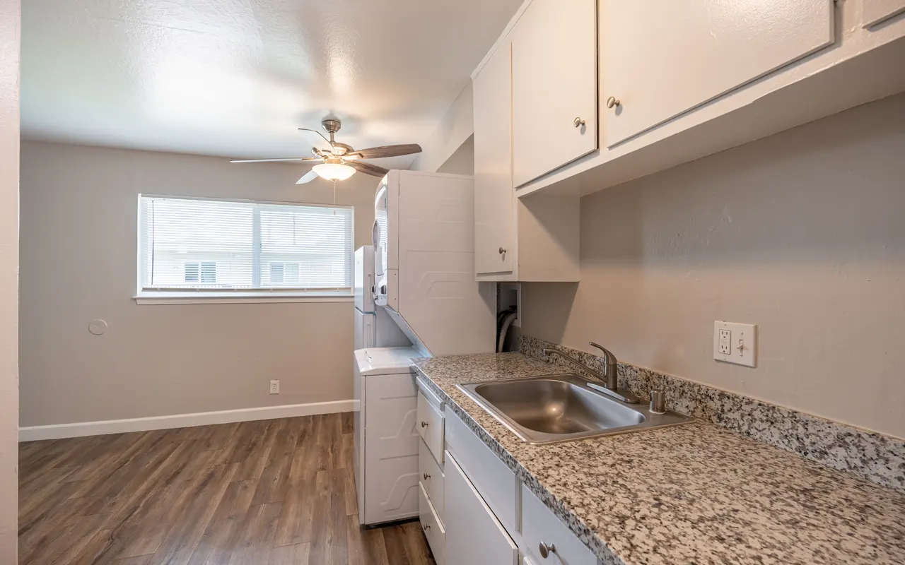 A clean, modern kitchen with white cabinetry, granite countertops, and a window letting in natural light. A ceiling fan is visible above the sink area, and the flooring is wood-like.