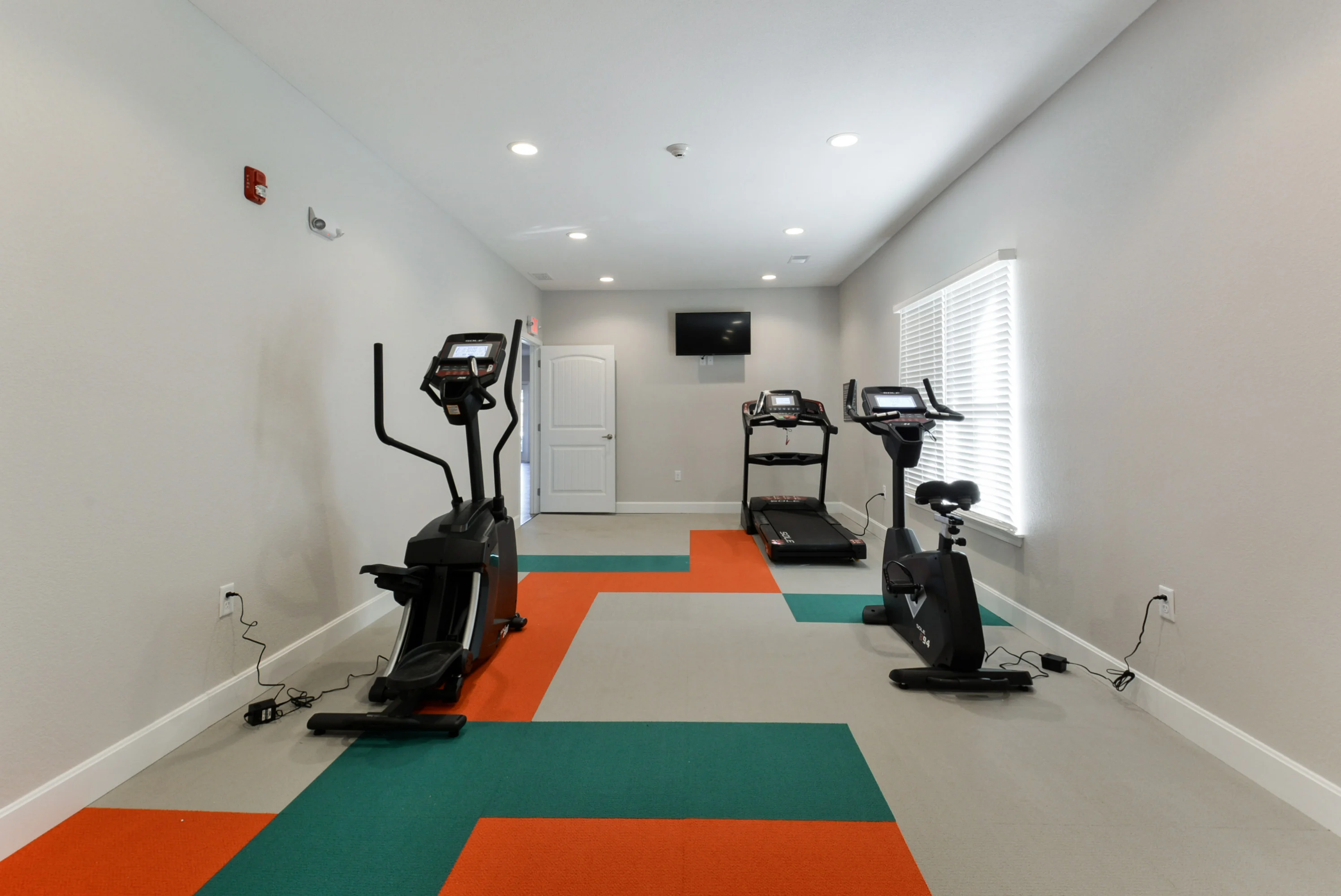A modern fitness room featuring an elliptical machine, a treadmill, and a stationary bike, with colorful flooring in teal, orange, and gray. Natural light comes from windows, and there is a wall-mounted TV.