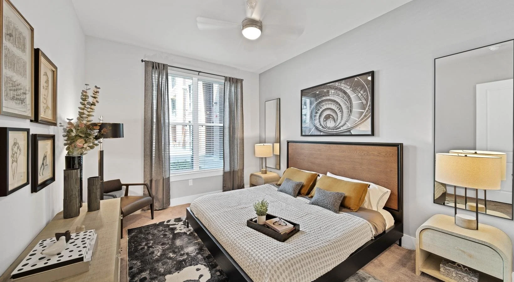 A stylish bedroom featuring a large bed with decorative pillows, a bedside table with a lamp, and artwork on the walls. The space has natural light coming through a window and a cozy rug on the floor.