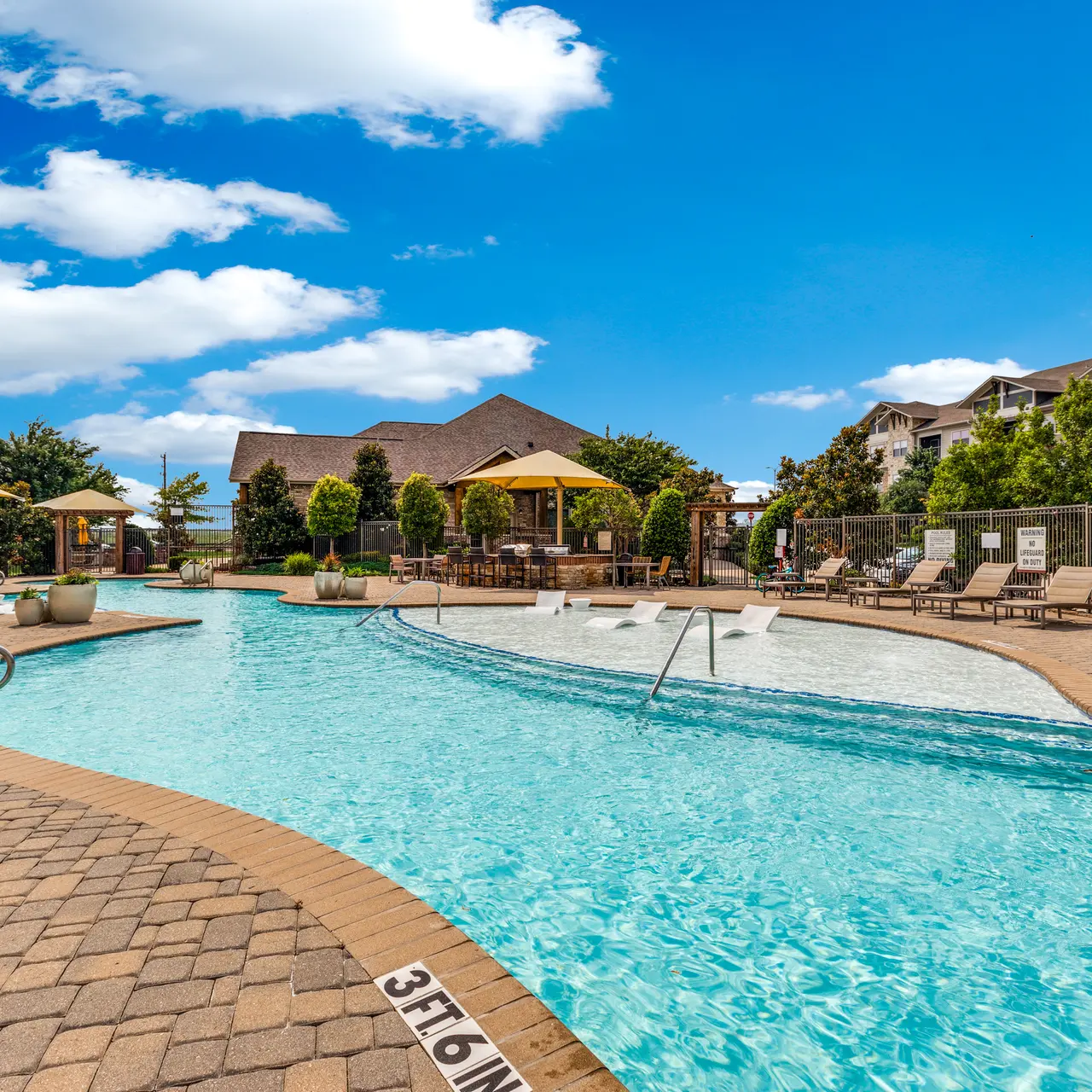 A sparkling blue pool surrounded by lounge chairs and umbrellas, set against a bright blue sky with fluffy white clouds. The area is well landscaped with trees and plants.