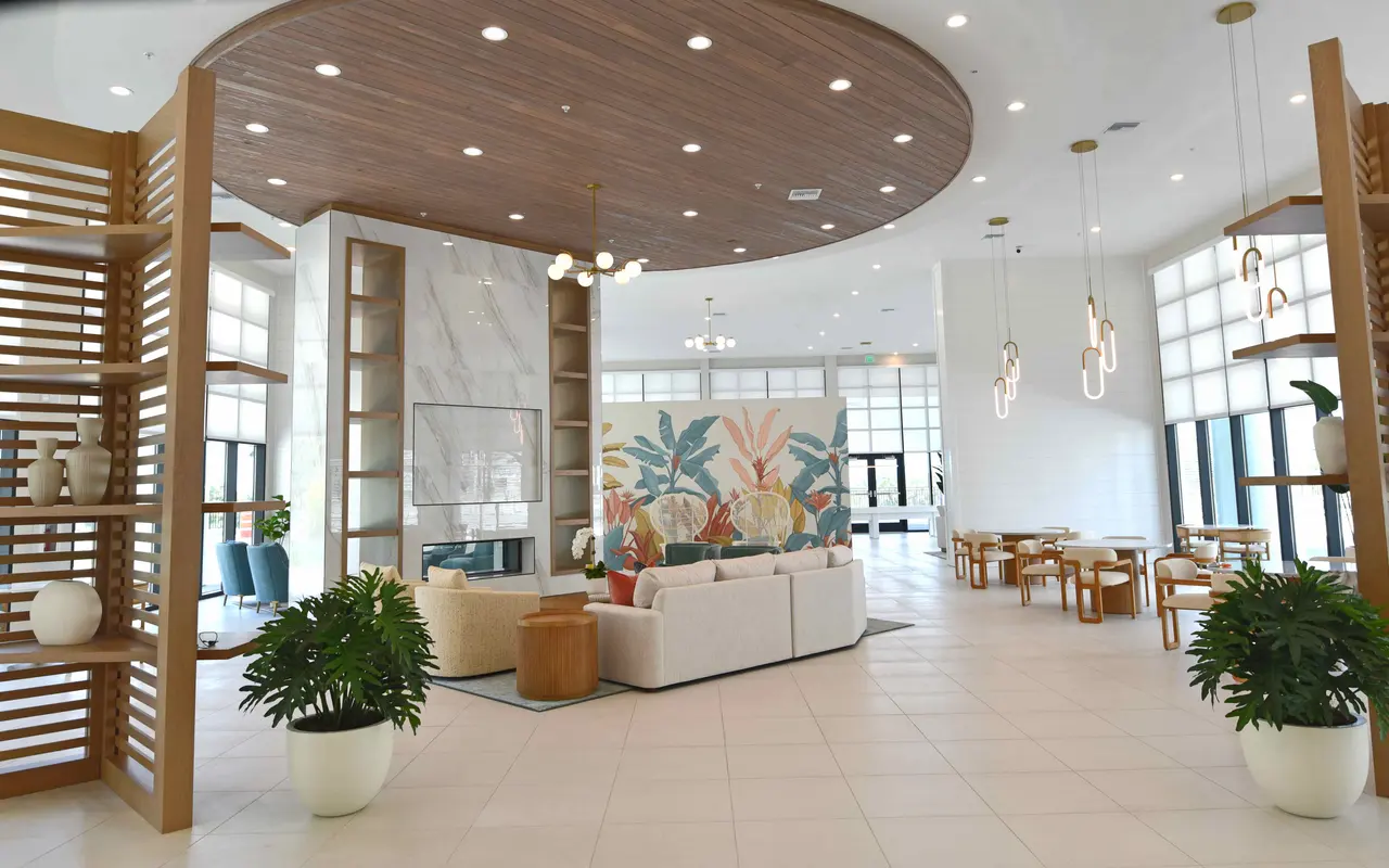 A spacious modern lobby area featuring a mix of natural wood elements and contemporary furnishings. There is a seating area with a large white sofa, a vibrant floral mural in the background, and wooden furniture arranged for social gatherings. Large windows allow natural light to flood the area, enhancing the open and airy atmosphere.