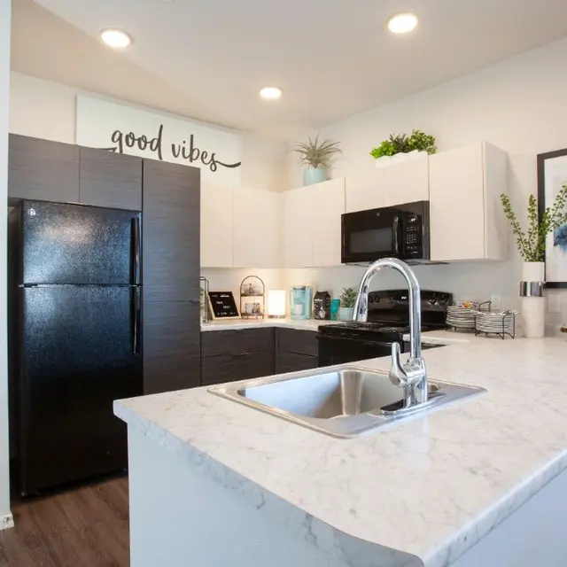 A modern kitchen featuring dark cabinetry, a black refrigerator, white countertops, and a stylish sink. The wall displays the phrase 'good vibes.'