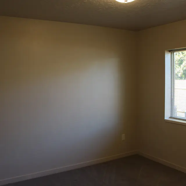An empty room with beige wall paint, a window with blinds letting in natural light, and a carpeted floor.