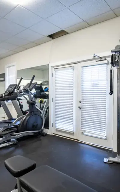 A well-lit gym interior featuring various exercise equipment including treadmills, an elliptical, and a weight machine. The floor is covered in a dark material, and there are large windows with blinds. A television is mounted on the wall.