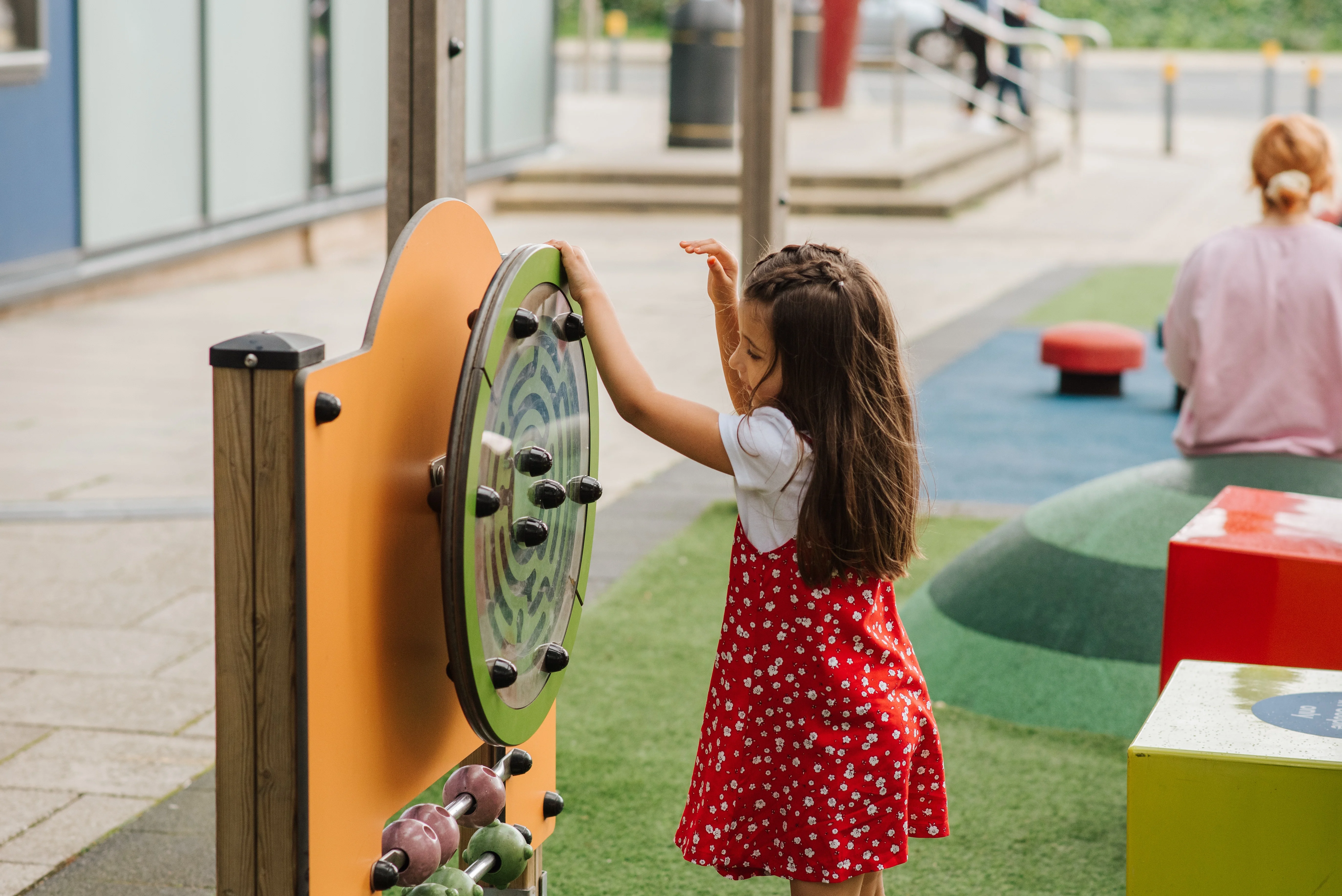 Child Playing at the Playground A young girl with long hair in a red dress with white polka dots is interacting with a colorful playground activity board. She is standing in front of the board which has spinning elements and is focused on the activity. In the background, other playground equipment and some people are visible.