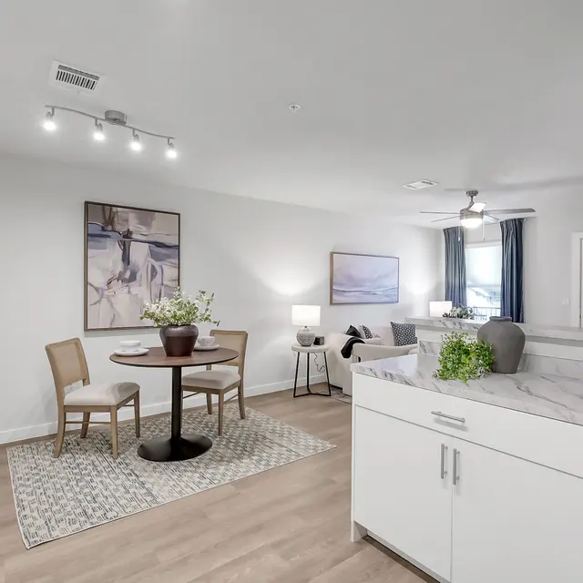 A bright and modern apartment interior featuring a small dining table with two chairs, a small potted plant on the table, a kitchen in the foreground, and a cozy living area with artwork on the walls and natural light from windows.