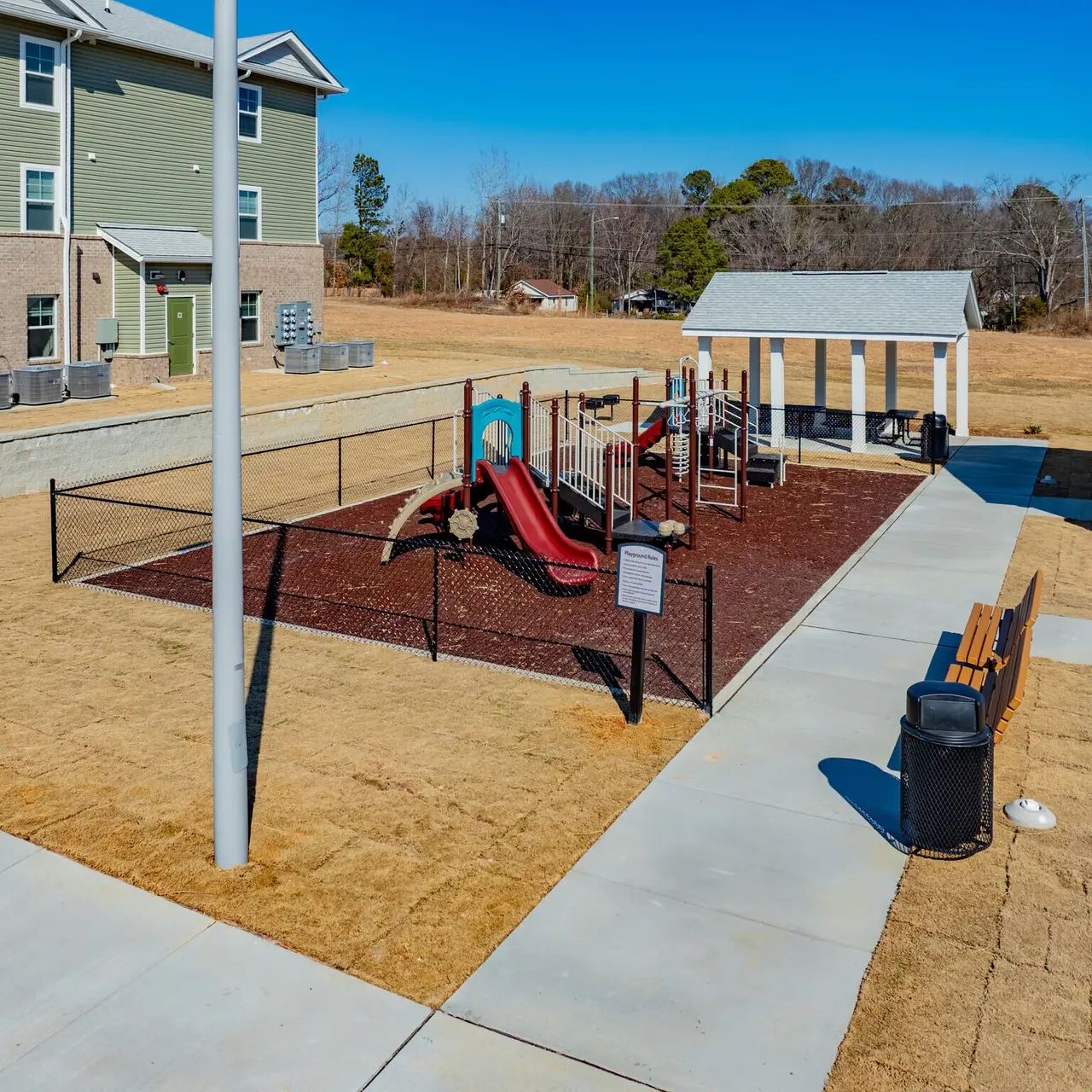 A playground with red slides and climbing structures enclosed by a fence, situated next to a grassy area and a few buildings in the background. There is also a pavilion with shelter and a bench nearby.