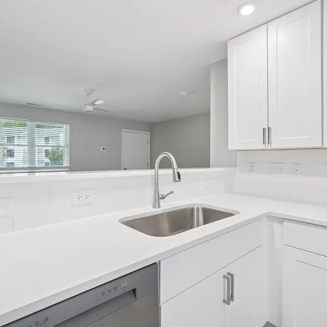 A modern kitchen featuring white cabinetry, a stainless steel sink, and a bright countertop. The background includes a glimpse of a living space with natural light coming from a window.