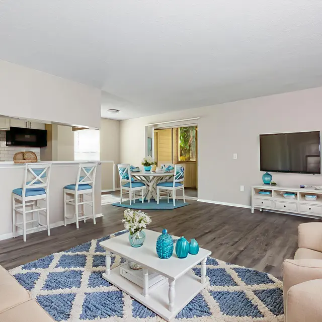 A modern living room featuring light-colored walls and a cozy seating arrangement with a beige sofa and decorative pillows. A coffee table with turquoise accents sits on a patterned rug. In the background, there's a dining area with a view of a kitchen and a TV mounted on the wall.