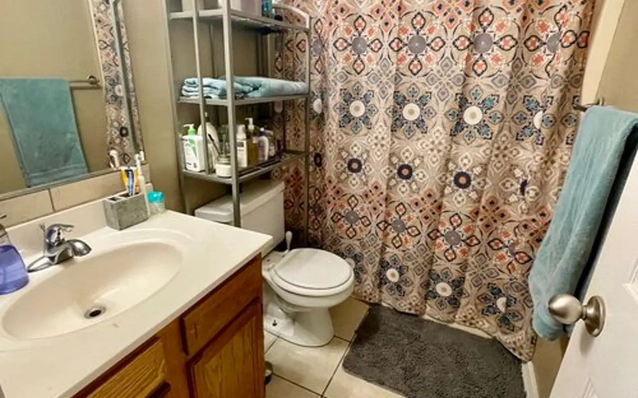 Compact Bathroom Interior A small bathroom featuring a beige wall, a white sink with wooden cabinets, and a shower curtain with a patterned design. A metal shelf stands next to the shower holding toiletries, and a light gray bath mat is on the floor.