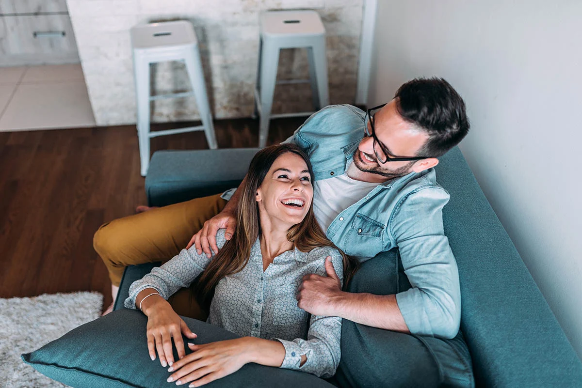A happy couple sitting on a couch, smiling and enjoying each other's company. The woman is leaning against cushions, while the man is playfully holding her shoulder. The background includes a light-colored wall and stools.