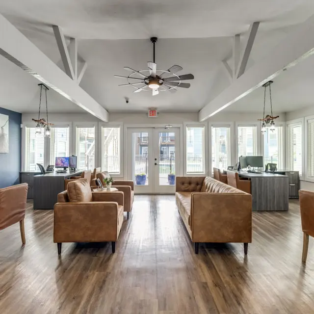 A spacious and modern lobby area with brown leather sofas, wooden flooring, and large windows allowing natural light. The space includes a ceiling fan and a reception area with desks, showcasing a welcoming atmosphere.