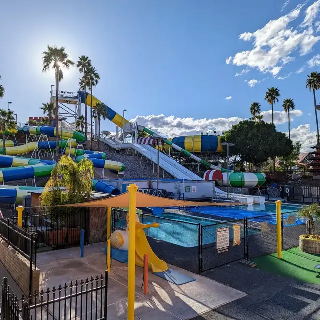 A colorful water park featuring a variety of large water slides surrounded by palm trees and an open sky with clouds. It includes a swimming pool and other water attractions.