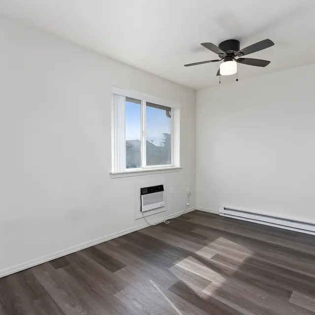 Empty Room with Ceiling Fan A vacant room with light-colored walls, wooden flooring, and a ceiling fan. Large window allowing natural light to enter.