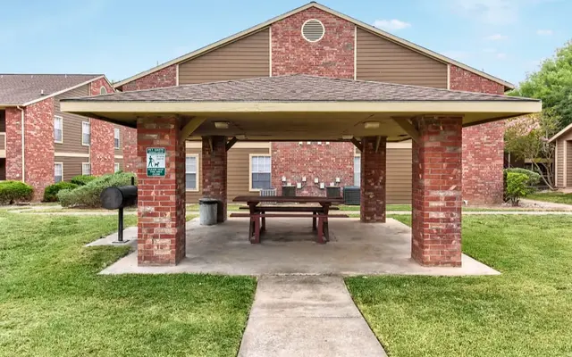 A brick pavilion with a picnic table underneath and a grassy area surrounding it, located near residential buildings.
