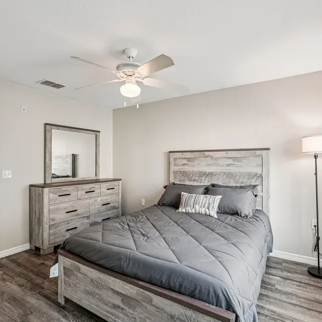 A well-lit modern bedroom featuring a wooden bed, nightstand, and dresser. The walls are painted in a light color and there is a lamp and ceiling fan present.