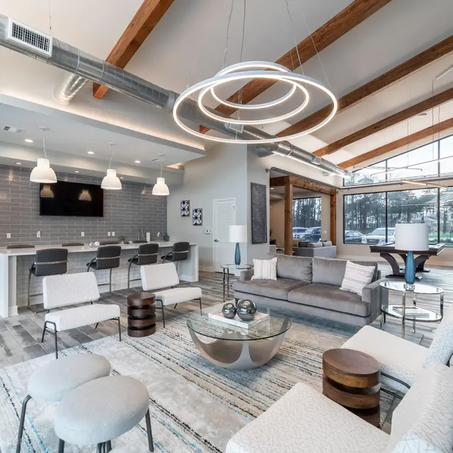 A spacious modern living room featuring a light color palette, large windows, and high wooden ceilings. The area includes a comfortable seating arrangement with a sofa, lounge chairs, and a coffee table. A kitchen bar with stools is seen in the background, along with modern lighting fixtures.