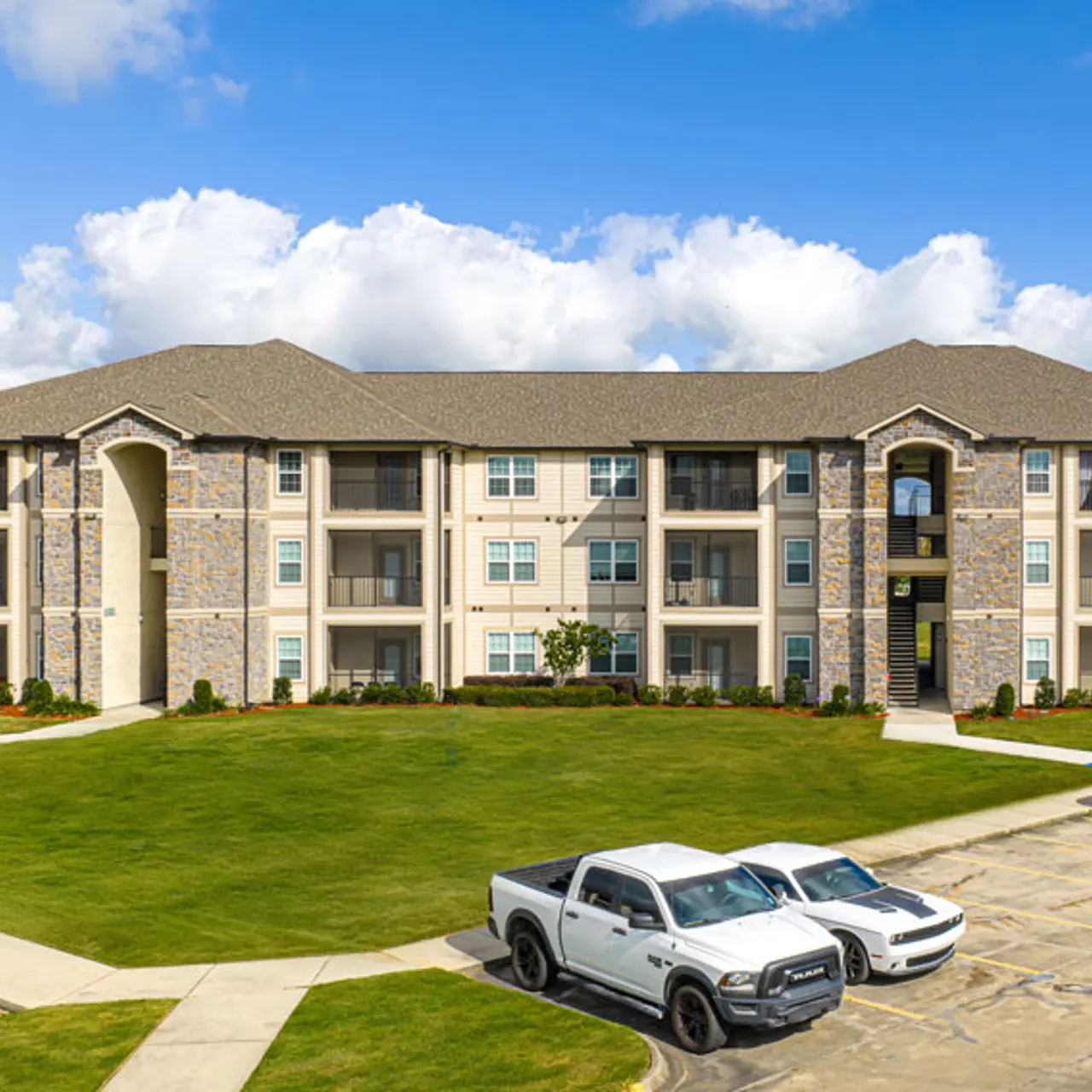 A modern three-story apartment complex with a stone and stucco exterior on a sunny day. The building has multiple balconies and a well-maintained green lawn in front, along with several parked vehicles.