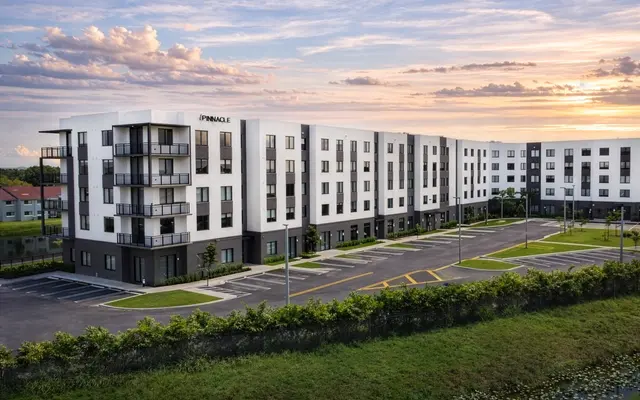 A modern apartment complex with a three-story building featuring balconies, surrounded by green lawn and a serene sky during sunset.