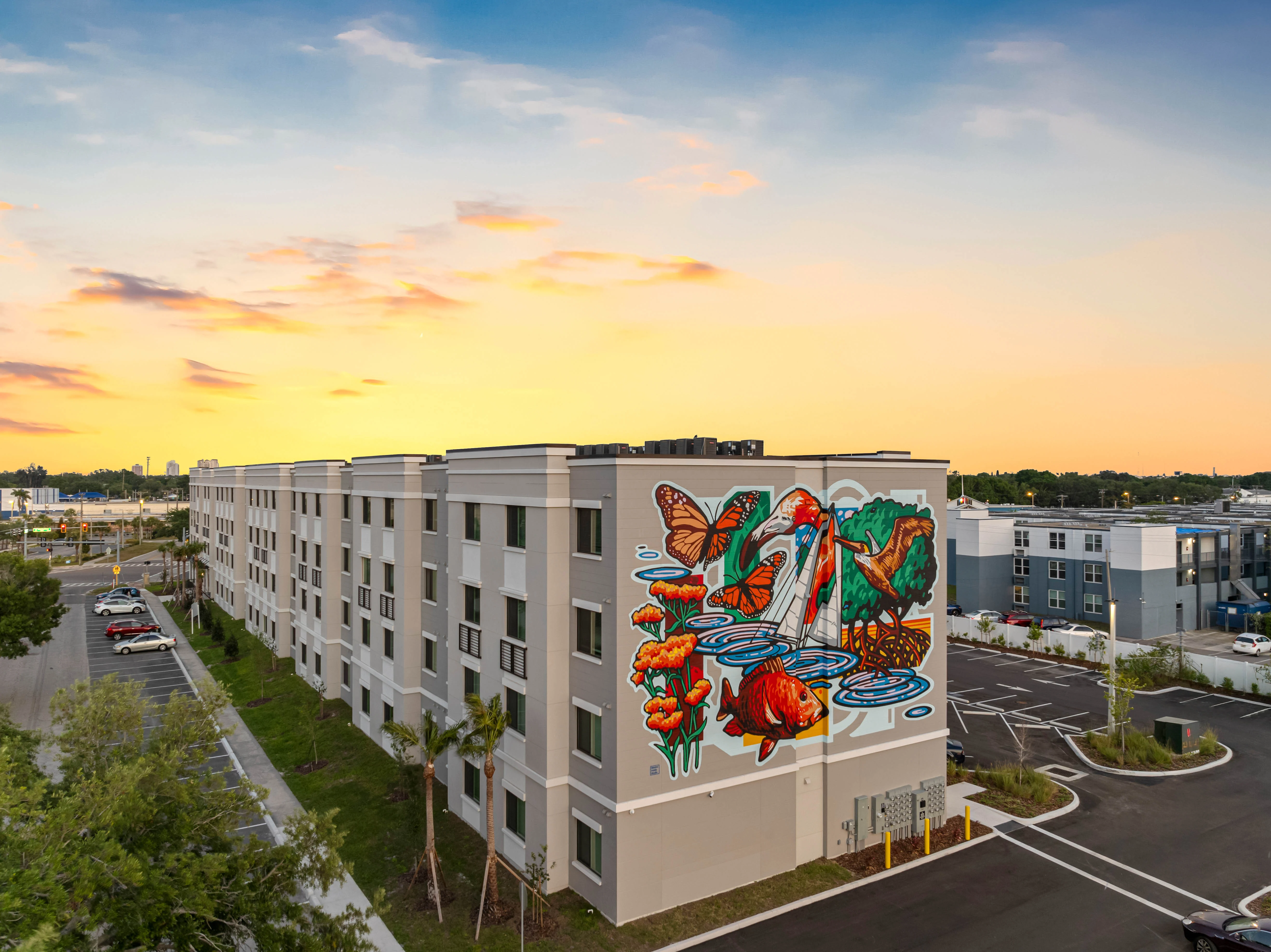A multi-story hotel featuring a vibrant mural of flowers and butterflies on one side, set against a sunset sky. The foreground includes parking spaces and palm trees.