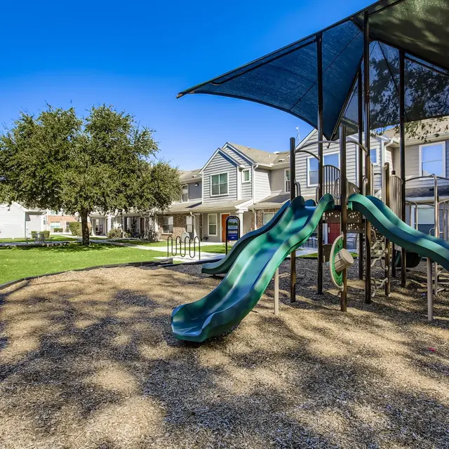 A playground with green slides surrounded by a sandy area and residential buildings in the background under a clear blue sky.