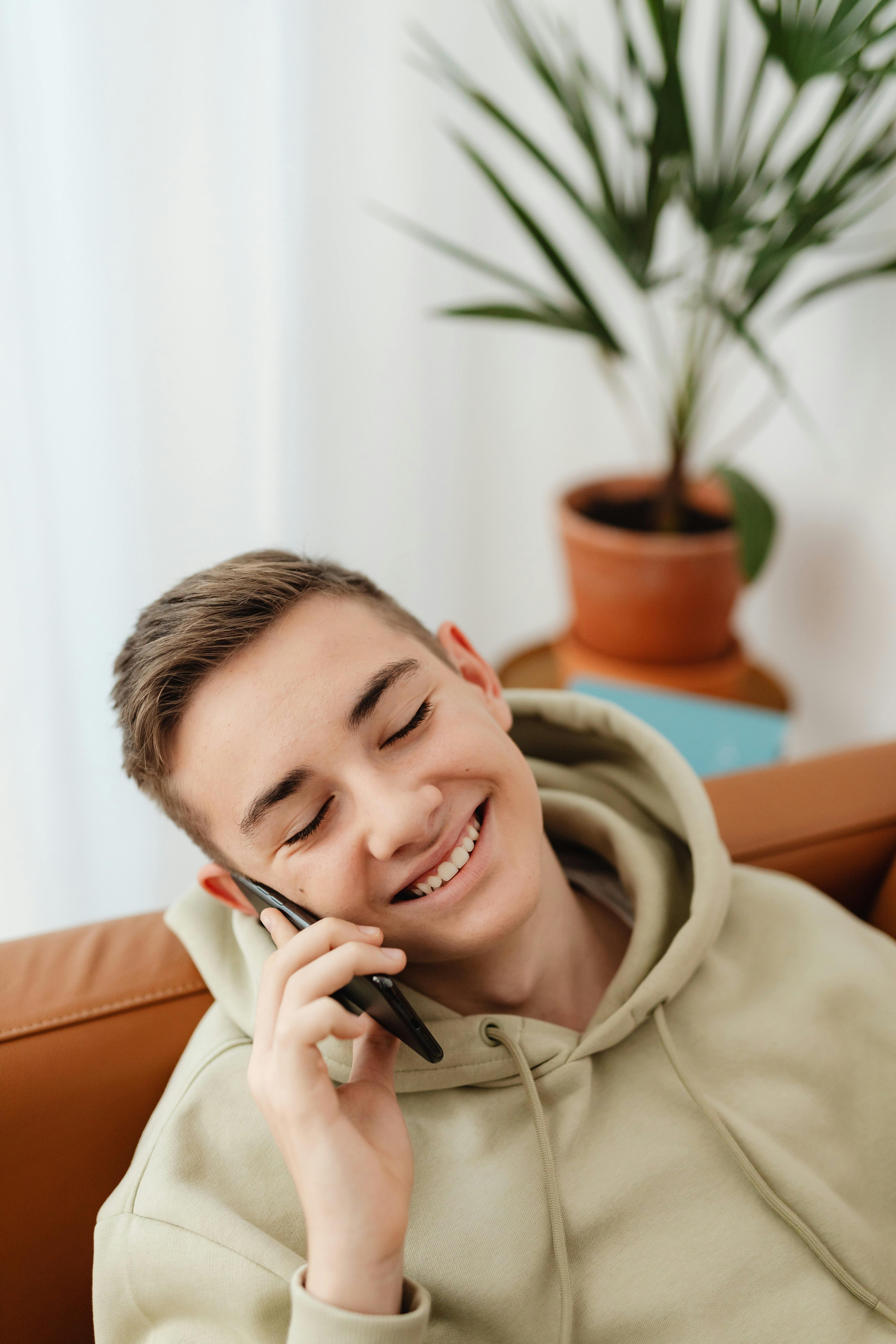 A young man with short light brown hair is smiling while talking on a mobile phone. He is wearing a light green hoodie and is seated on a brown couch. A potted plant is visible in the background.