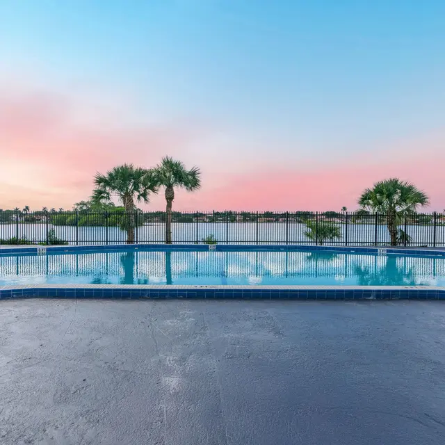 A tranquil swimming pool overlooking a scenic lake at sunset, with palm trees and a soft pink and blue sky.