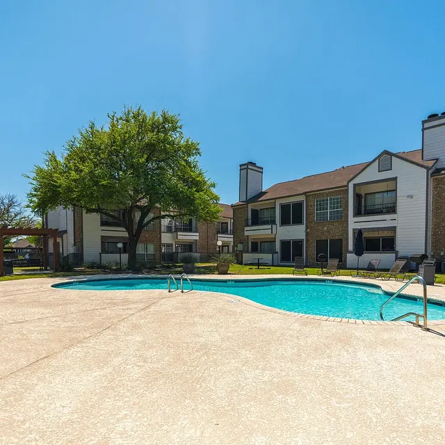 A sunlit apartment complex featuring a swimming pool surrounded by green grass and a tree, with residential buildings in the background.