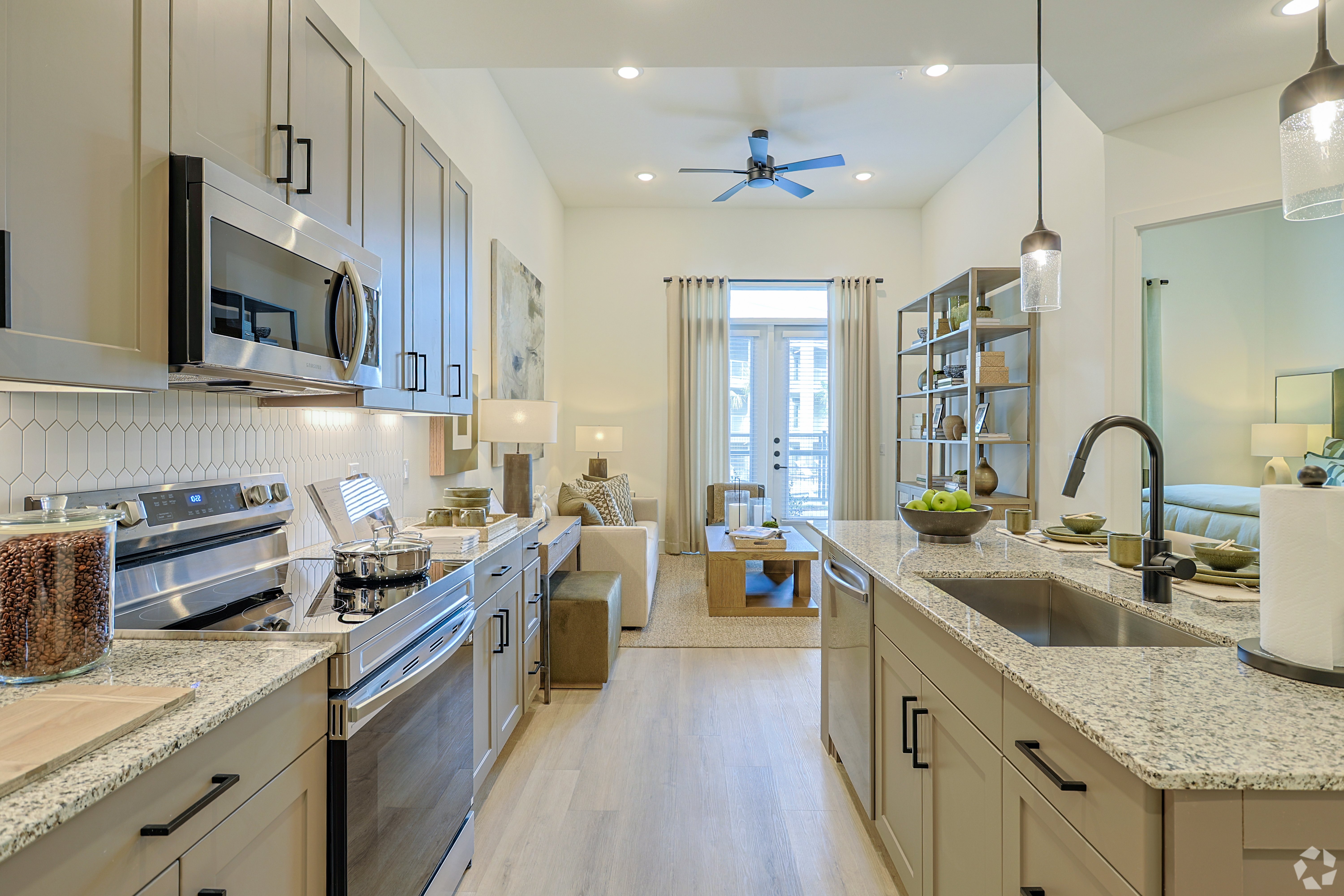 Modern Kitchen and Living Room Design A modern kitchen with stainless steel appliances and granite countertops opens up to a bright living area with light-colored furniture and a balcony in the background.