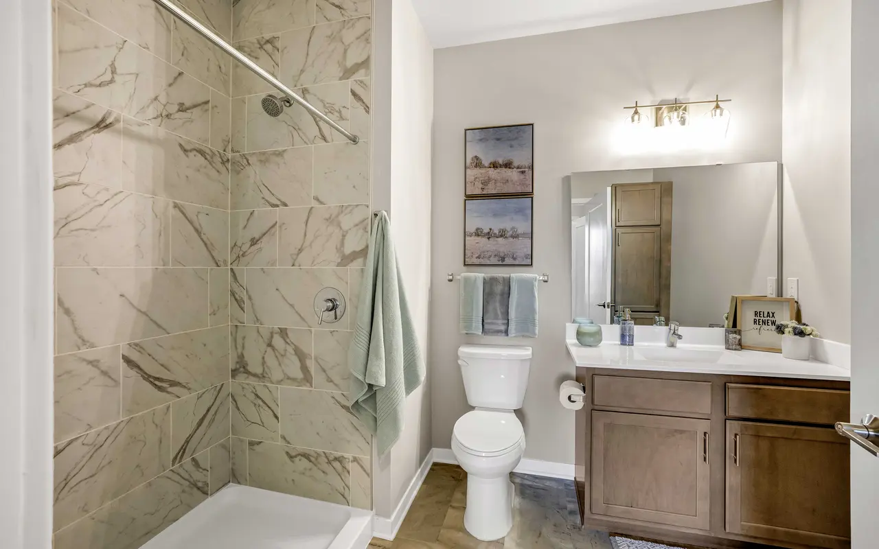 A modern bathroom featuring a walk-in shower with marble-style tiles, a white toilet, and a wooden vanity with a mirror. The space is well-lit and includes a towel rack and decorative wall art.