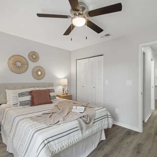 A cozy bedroom featuring a queen-sized bed with striped bedding, an end table with a lamp, and decorative wall hangings. The room has light gray walls and a ceiling fan. A doorway leads to another area in the background, suggesting an open layout.