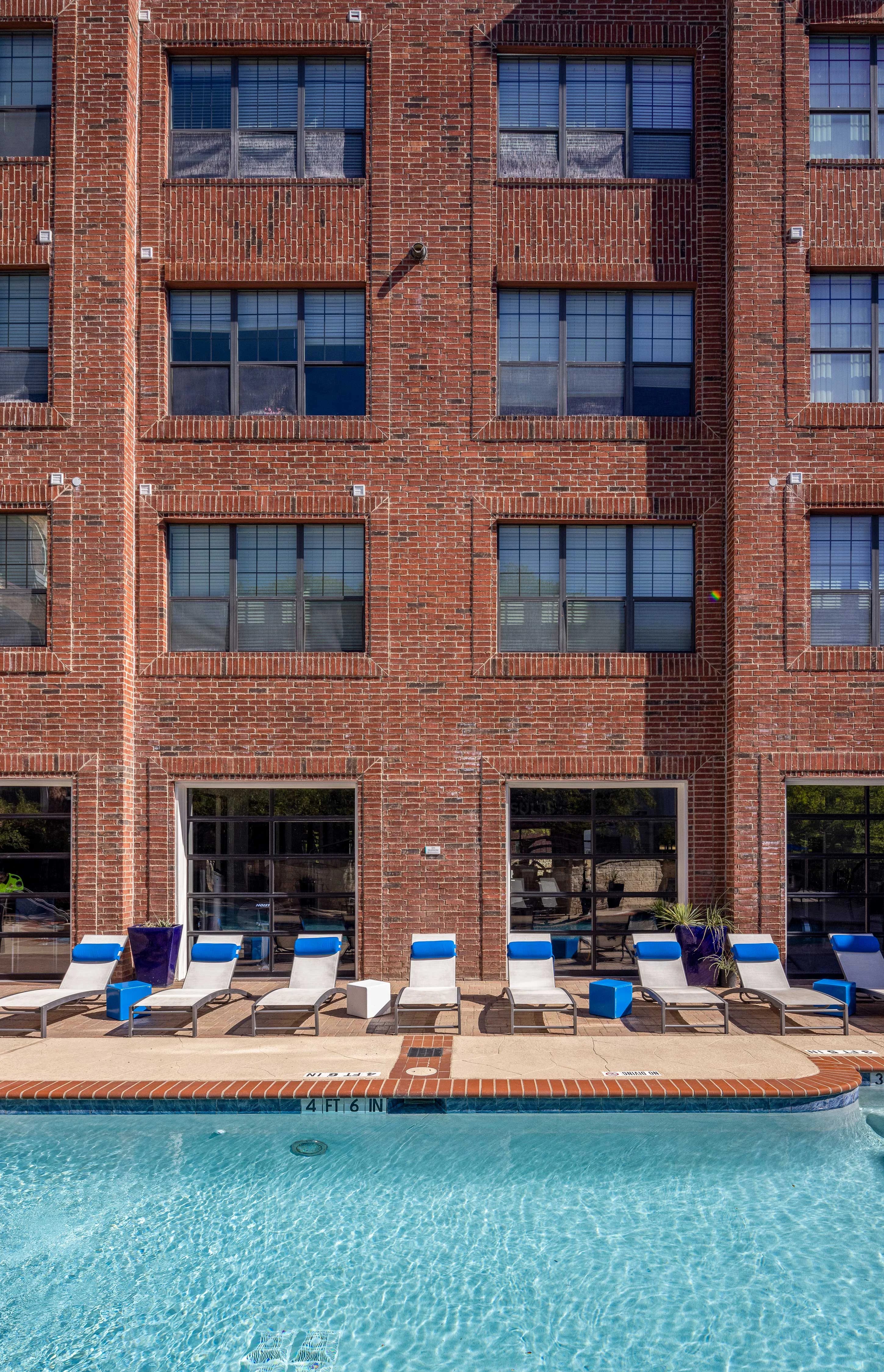 View of a swimming pool in front of a brick building, with lounge chairs lined up by the poolside.