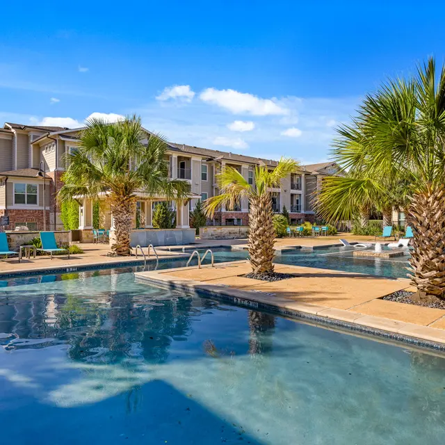 A spacious outdoor pool area of a luxury apartment complex, featuring palm trees and lounge chairs under a clear blue sky.