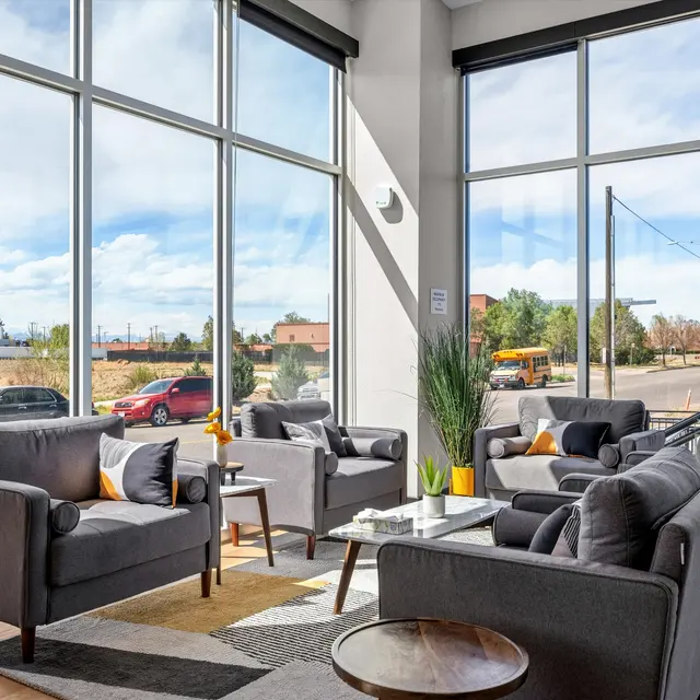 A modern lounge area with large windows allowing natural light, featuring gray armchairs with yellow cushions and a glass coffee table, plants add decor to the space, and a view of vehicles outside.