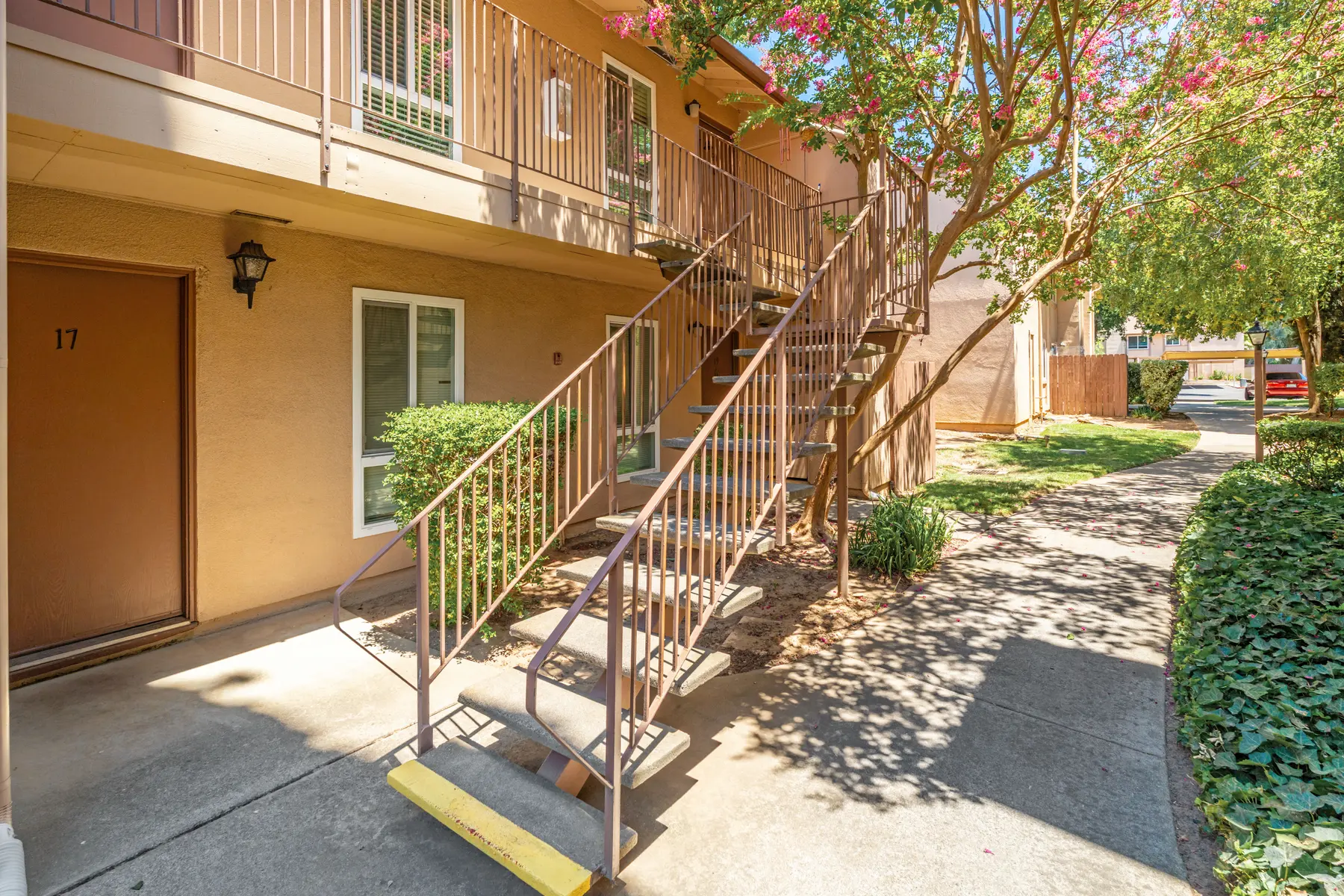 Exterior view of an apartment building with stairs leading to upper floors, surrounded by greenery and a paved walkway.