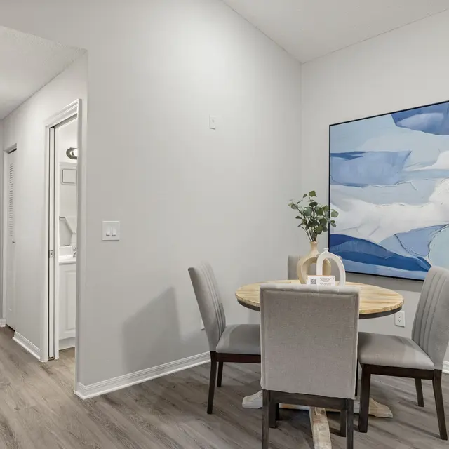 A modern dining area featuring a round table with four chairs, a simple vase with greenery, and a large abstract painting on the wall. The decor is minimalist, with light wood flooring and soft neutral walls. A doorway leads to another room with windows allowing natural light to enter.