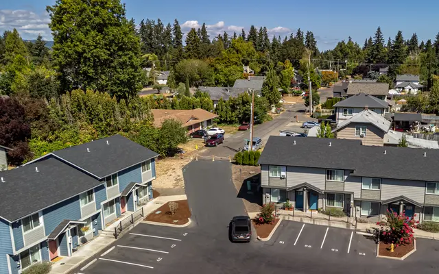Aerial view of a residential area with trees, houses and parked cars. The foreground shows a parking lot with two-story buildings, and the background features a mix of greenery and homes.