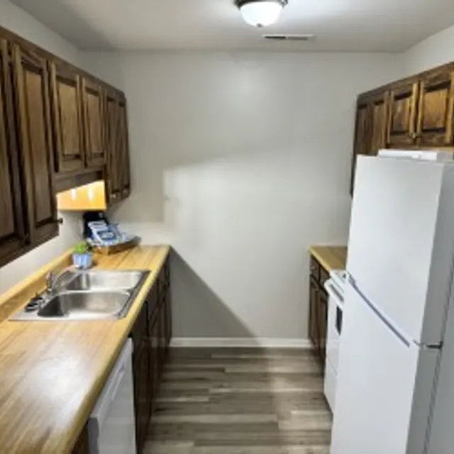 A small kitchen with wooden cabinets and light brown countertops. A white refrigerator is on the right, and a sink with a silver faucet is visible on the left. The floor is wooden and the walls are painted light. An emergency fire extinguisher is mounted near the refrigerator.