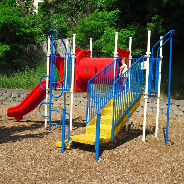 Colorful Playground Adventure A colorful playground with a red slide and yellow stairs. A child is playing on the equipment with blue railings. The background features greenery and a stone wall.