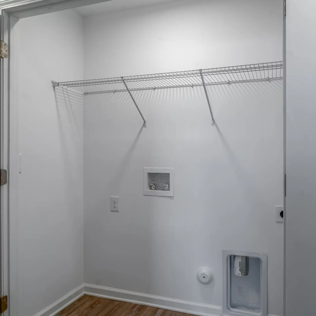 A view of a small, empty laundry room with a wire shelf, light-colored walls, and wooden flooring.