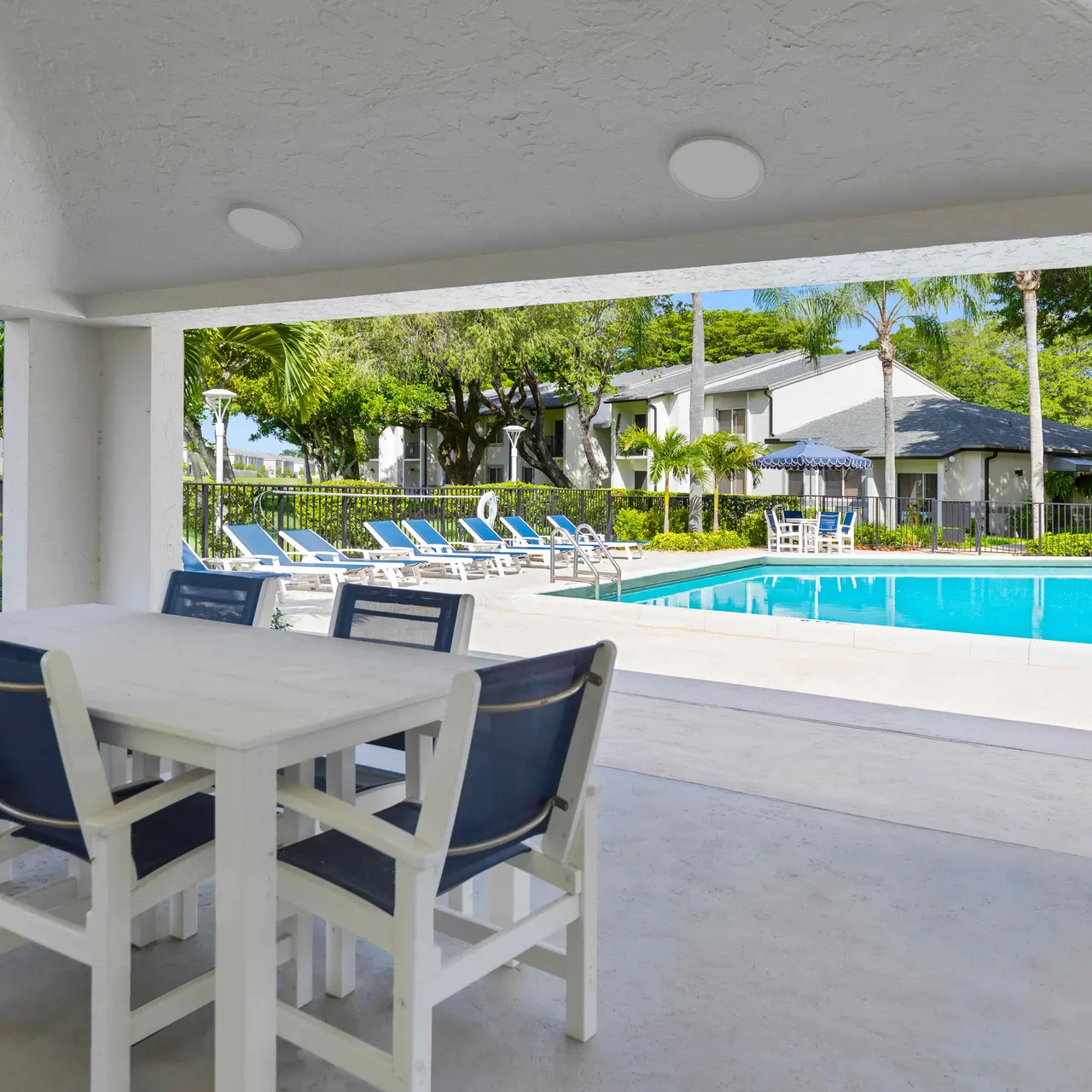 Outdoor Poolside Community Area View of a pool area from an outdoor dining space with a table and chairs, surrounded by lounge chairs and greenery.