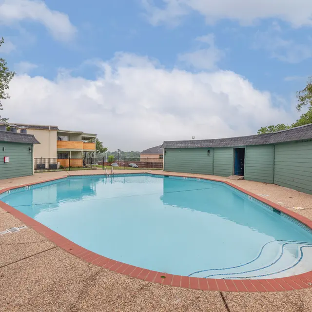The Hills - A clear swimming pool surrounded by light-colored pavement and two green storage buildings. The background features apartment buildings and a cloudy sky.
