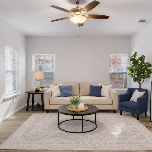A cozy living room with a beige couch, blue pillows, a black coffee table, and a floor lamp. A plant is in the corner, and a sign that says 'Welcome' is displayed.